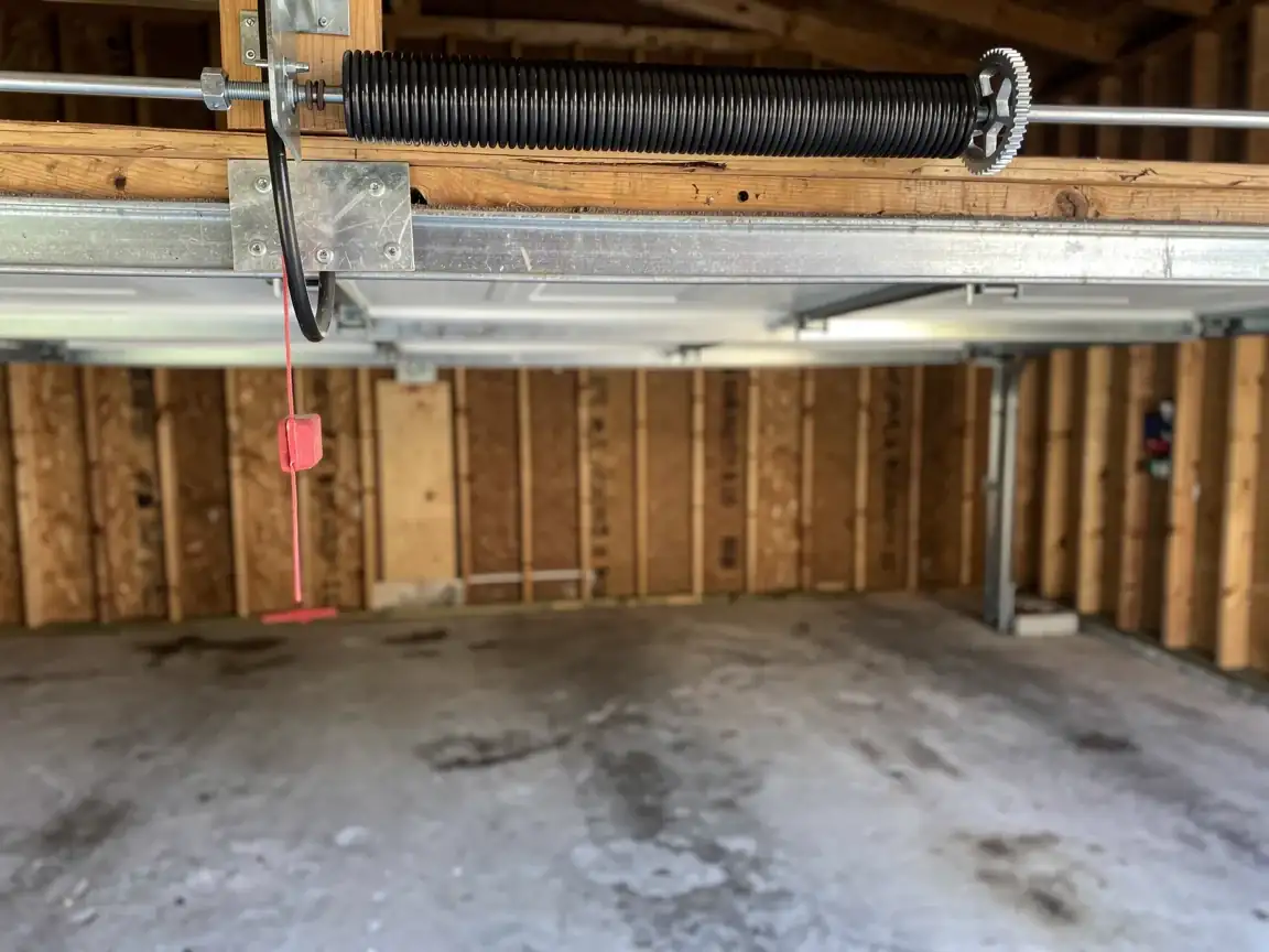 A black torsion spring is mounted above a white garage door in an empty garage.